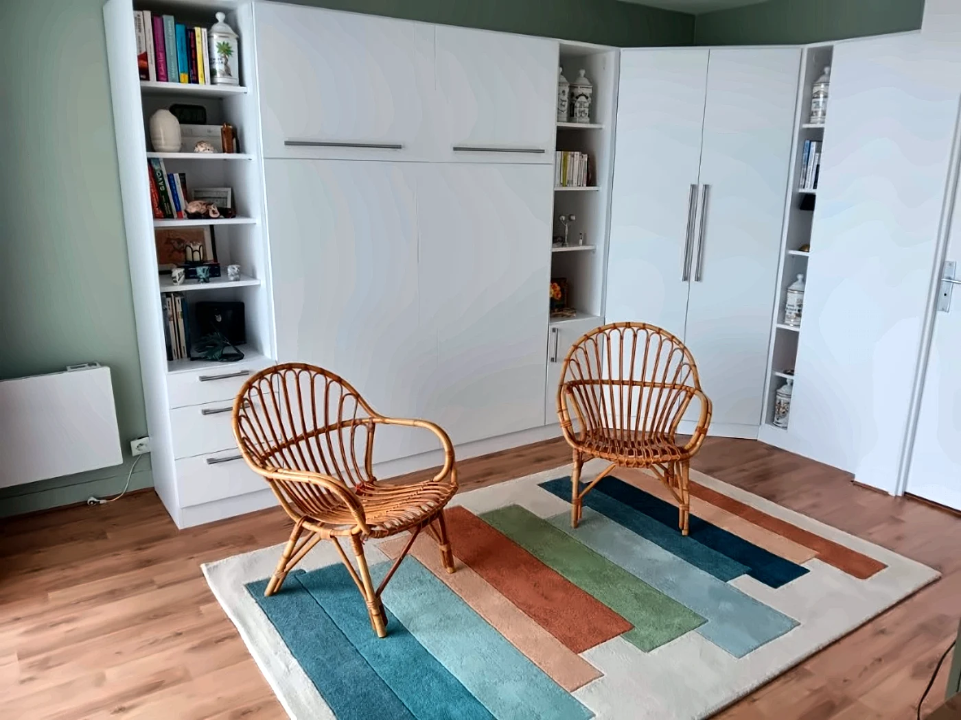 Coin salon avec deux fauteuils en rotin devant un lit escamotable fermé, bibliothèques et armoire d'angle blanches