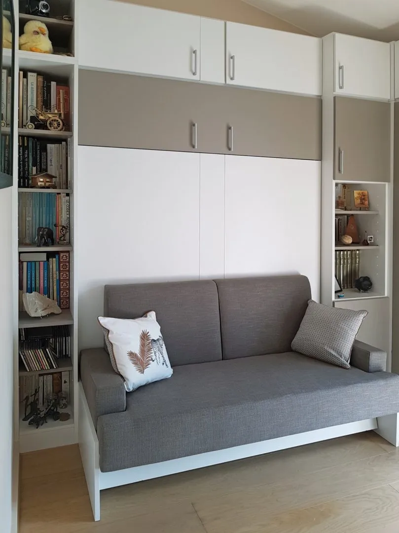 Lit escamotable fermé avec canapé gris, encadré de bibliothèques et de placards hauts